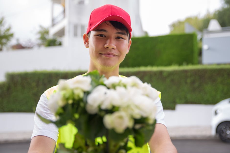 A delivery person wearing a red cap and reflective vest is smiling and holding a bouquet of fresh white flowers with green foliage, wrapped in clear cellophane. The flowers appear to be roses or simil