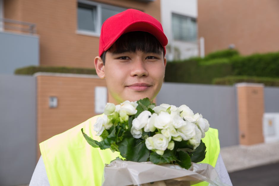 A young delivery person wearing a red cap and a high-visibility yellow vest is standing outdoors in front of modern residential buildings, holding a bouquet of fresh white damask roses wrapped in whit