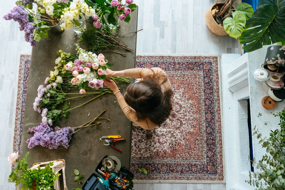 A florist with shoulder-length dark hair arranging a bouquet of pink and white roses, lisianthus, and green foliage on a rustic grey table indoors. The flowers are fresh with vibrant petals and sturdy