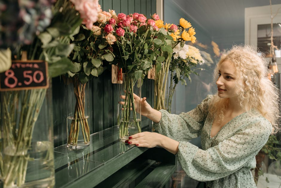 A woman with curly blonde hair wearing a light, floral-patterned dress arranging a bouquet of mixed fresh flowers in a glass vase at a florist shop. The bouquet includes pink roses and yellow chrysant