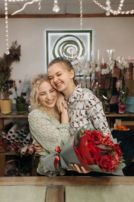 Two young women are smiling and hugging inside a floral shop, with one presenting the other a vibrant bouquet of red roses and red hypericum berries wrapped in gray paper with a red ribbon. The floral